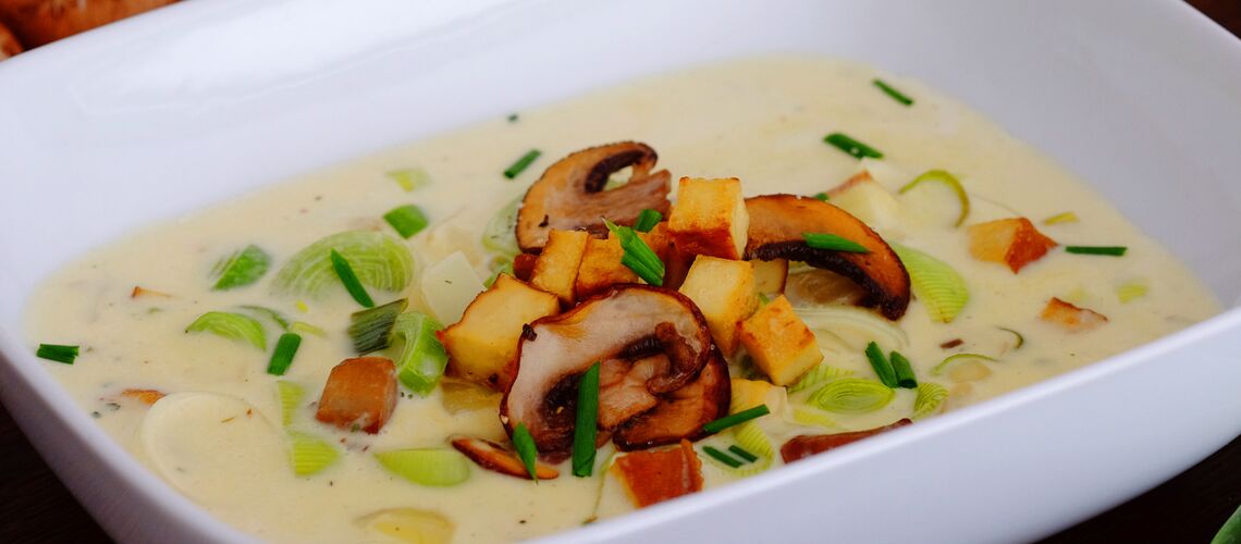 Eine helle Suppe mit Lauchringen und Champignon-Scheiben in einer weißen, eckigen Schüssel.