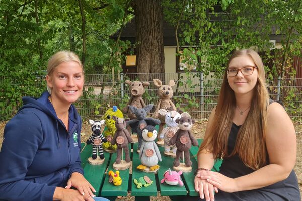 Zwei junge Frauen sitzen draußen an einem grünen Holztisch, auf dem diverse gehäkelte Stofftiere stehen.
