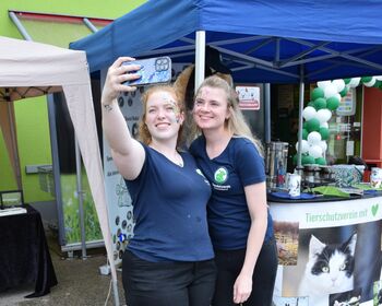 Zwei junge Frauen machen vor einem Infostand ein Selfie.