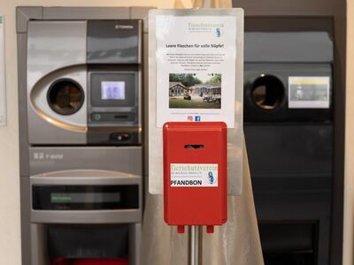 Ein roter Pfandbon-Behälter der auf einem Aluminiumpfahl vor zwei grauen Leergut-Automaten steht.
