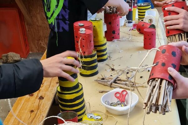 Ein langer Holztisch auf dem rot und schwarz-gelb bemalte Konservendosen stehen, die von Kindern mit Schnüren versehen werden.