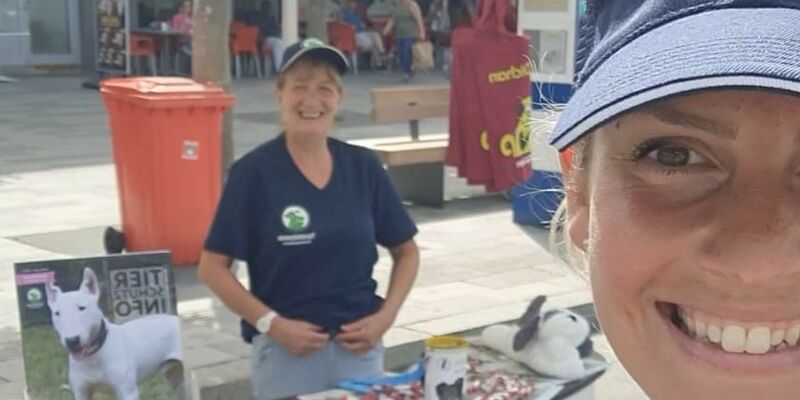 Eine Frau mit Schirmmütze mit Tierschutzverein-Logo lächelt im Vordergrund in die Kamera. Hinten eine weitere Frau hinter weißer Infostad-Theke.