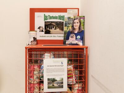 Ein roter Drahtkorb mit Futterpackungen darin. Oben auf dem Korb stehen Magazine der "Tierschutz-Info" und eine Spendendose.