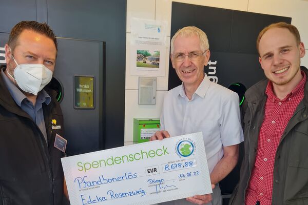 Drei Männer stehen vor einem Leergutautomaten und halten einen großen symbolischen Spendenscheck in die Kamera.