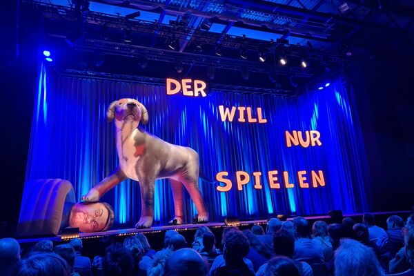 Vor dem noch geschlossenen Bühnenvorhang steht in blauem Lichtein riesiger aufgeblasener Plastikhund, der eine Pfote auf  den Kopf eines liegenden aufblasbaren Plastik-Menschen hält.