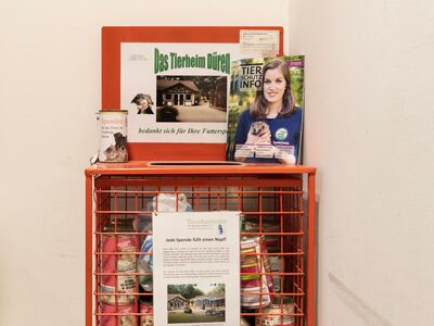 Ein roter Drahtkorb mit Futterpackungen darin. Oben auf dem Korb stehen Magazine der "Tierschutz-Info" und eine Spendendose.