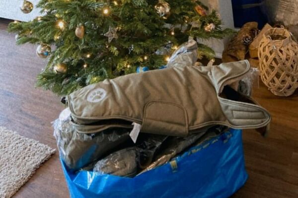 Eine große blaue Kunststofftasche, randvoll gefüllt mit neuen, teils verpackten Hundemänteln steht auf einem Holzfußboden vor einem festlich geschmückten Weihnachtsbaum.