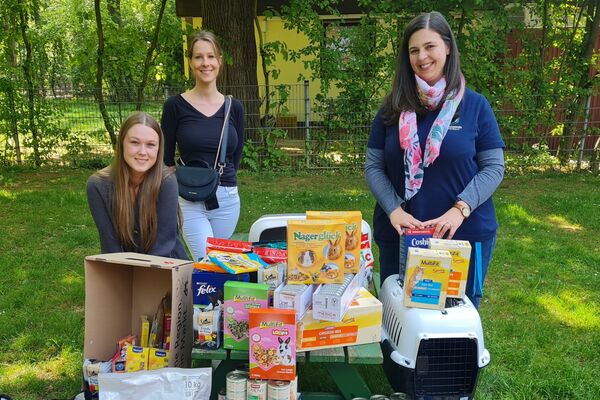 Drei Frauen stehen draußen hinter einem Tisch, auf dem Tierfutter und -Zubehör steht.
