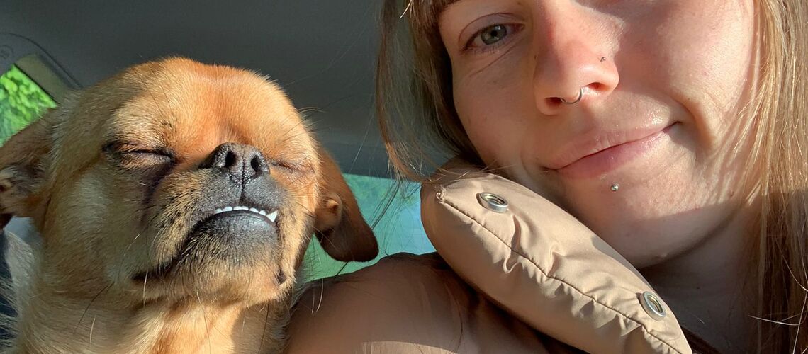 Eine Frau angeschnallt im Auto, an ihre Schulter lehnt sich mit geschlossenen Augen der kleine Hund Luke..