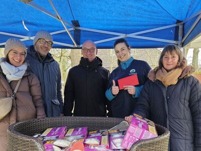 Fünf Personen in Winterkleidung steht draußen unter einem blauen Pavillondach. Davor auf einem grünen Tisch ein Kunststoff-Hundekorb voll pinkfarbener Futtertüten. Eine Frau hält einen roten Umschlag in die Höhe.