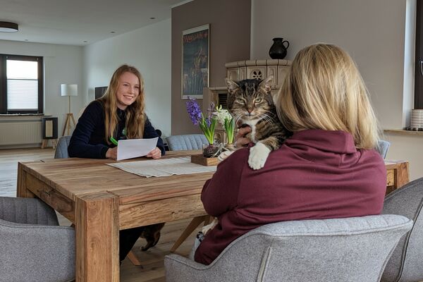Zwei Frauen sitzen an einem Holztisch. Eine der beiden in Tierheim-Kleidung macht sich Notizen, der andere Frau, die von hinten sichtbar ist, schaut eine graugetigerte Katze über die Schulter.