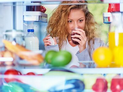 Eine junge Frau schaut in den geöffneten Kühlschrank mit Lebensmitteln hinein und riecht an einem Glas Marmelade.
