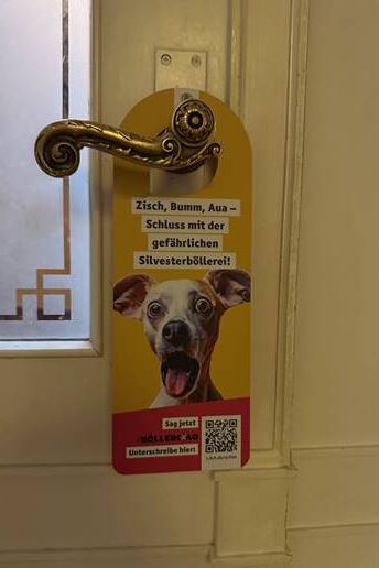 Ein Türaufhänger mit Aufschrift und dem Foto eines erschrockenen Hundes mit aufgesperrtem Maul und aufgerissenen Augen.