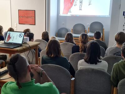 Eine Gruppe Kinder und Jugendlicher sitzt im Schulungsraum vor einer Leinwand-Präsentation.