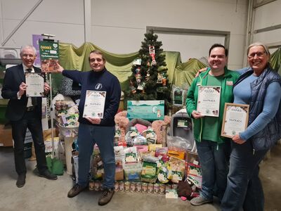 Vier Personen mit je einer Urkunde in den Händen stehen vor diversen Futter- und Sachspenden und sowie einem Weihnachtsbaum im Hintergrund.