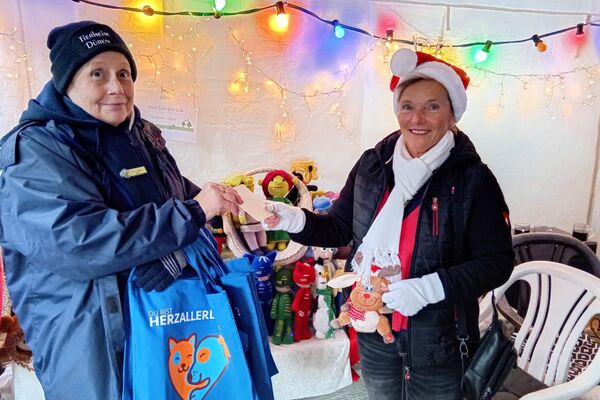 Zwei Frauen, wovon eine mit Weihnachtsmann-Mütze, halten einen Umschlag in den Händen. Darüber hängt im Pavillion eine bunte Lichterkette und im Hintergrund stehen Häkeltiere auf einem Tisch.
