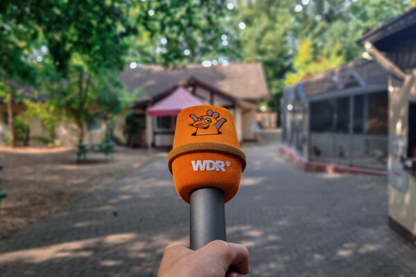 Ein oranges Mikrofon mit dem Bild der Maus sowie den Aufdruck "WDR" wird vor die Kamera gehalten. Dahinter leicht verschwommen ein Weg mit Volieren und dem Tierheim-Büro.