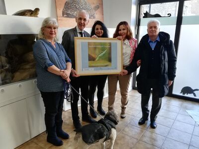 Drei Frauen und zwei Männer stehen in einer Reihe und halten ein Bild hoch. Davor ein mittelgroßer Hund mit Hundemantel.