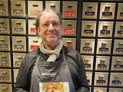 Ein Mann in grauer Schürze steht vor einer Wand mit vielen kleinen Teeschubladen und hält das vegane Kochbuch in der Hand.