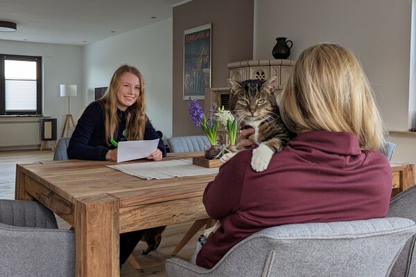 Zwei Frauen sitzen an einem Holztisch. Eine der beiden in Tierheim-Kleidung macht sich Notizen, der andere Frau, die von hinten sichtbar ist, schaut eine graugetigerte Katze über die Schulter.